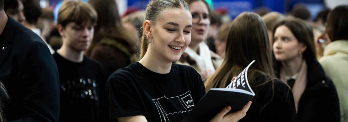 Jauna moteris su surištais plaukais ir juodais marškinėliais, ant kurių yra „KTU“ logotipas, šypsosi žiūrėdama į atverstą brošiūrą ar žurnalą. Aplink ją – būrys žmonių, daugelis jų taip pat jauni, lyg dalyvautų renginyje ar mugėje.
