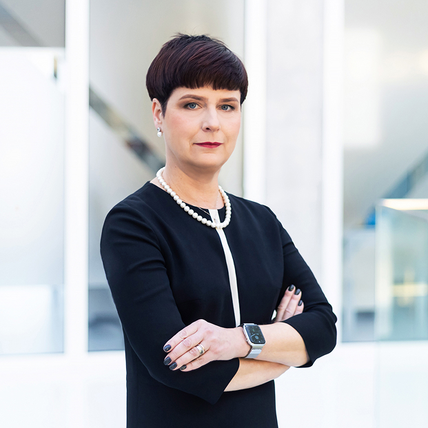 KTU strateginių partnerysčių prorektorė prof. dr. Edita Gimžauskienė KTU strateginių partnerysčių prorektorės portretinė nuotrauka. Moteris trumpais tamsiais plaukais vilki tamsų drabužių derinį ir perlų vėrinį. Ji stovi sukryžiavusi rankas ir žvelgia tiesiai į kamerą šviesioje universiteto aplinkoje.