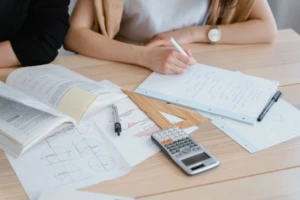 Studentė sprendžia matematikos užduotis – ant stalo išdėlioti vadovėliai, sąsiuviniai, liniuotės, skriestuvas ir skaičiuotuvas