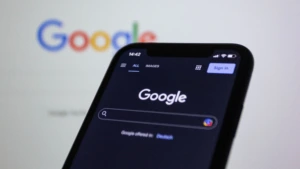 Išmaniojo telefono ekrane atidaryta „Google“ paieškos sistema, fone matomas neryškus „Google“ logotipas