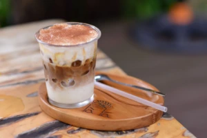 &Scaron;alta latte kava su grietinėle, patiekta ant kaimi&scaron;ko medinio stalo jaukioje aplinkoje